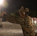 A military service member in digital camouflage uniform is firing a pistol at a nighttime shooting range. The shooter is using a two-handed grip stance with arms extended, wearing hearing protection and eye protection. The scene is illuminated by artificial range lighting against a dark background.