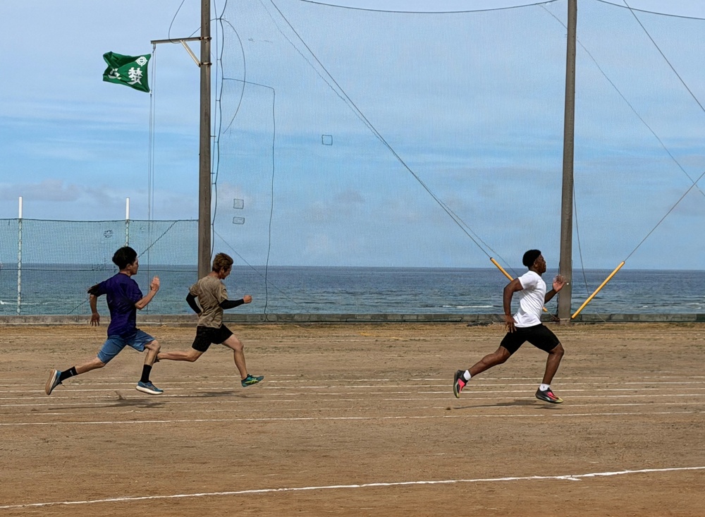 Three young athletes are running on a dirt track near a coastal location, with one runner in the lead holding what appears to be a yellow javelin or pole. A large net structure stands behind them, likely a baseball or sports backstop, with the ocean visible in the background. A green flag with Japanese characters is flying from a pole in the upper left area.