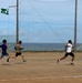 Three young athletes are running on a dirt track near a coastal location, with one runner in the lead holding what appears to be a yellow javelin or pole. A large net structure stands behind them, likely a baseball or sports backstop, with the ocean visible in the background. A green flag with Japanese characters is flying from a pole in the upper left area.