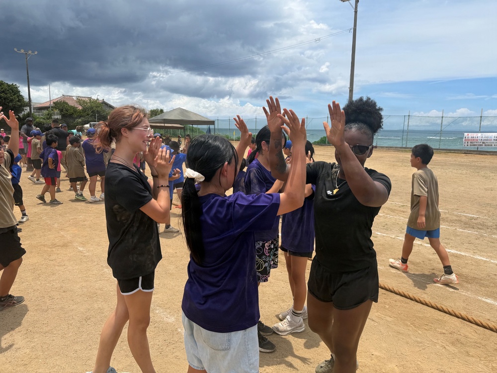 A group of children and young adults are participating in an outdoor sports day or field event on a dirt athletic field near a coastline. Participants are engaged in high-fiving and celebratory gestures, suggesting a team activity or game conclusion. The setting appears to be in East Asia, likely Japan, based on visible signage and architectural elements in the background.
