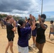 A group of children and young adults are participating in an outdoor sports day or field event on a dirt athletic field near a coastline. Participants are engaged in high-fiving and celebratory gestures, suggesting a team activity or game conclusion. The setting appears to be in East Asia, likely Japan, based on visible signage and architectural elements in the background.