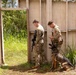 Two military personnel in MultiCam/OCP camouflage uniforms are conducting what appears to be a training exercise at a mock structure or shoot house. One soldier carries an assault rifle while the other holds a device (likely a phone or tablet) and handles a military working dog (MWD) in a tactical vest. The setting suggests a K9 handler training scenario at a military training facility.