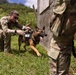 Two military personnel in MultiCam/OCP camouflage uniforms are working with a military working dog (MWD) wearing a tactical vest in a lush, green outdoor training environment. One soldier is crouched down handling the dog, which appears alert and energetic with its mouth open. The setting appears to be a field training exercise near a wooden structure with mountainous terrain visible in the background.