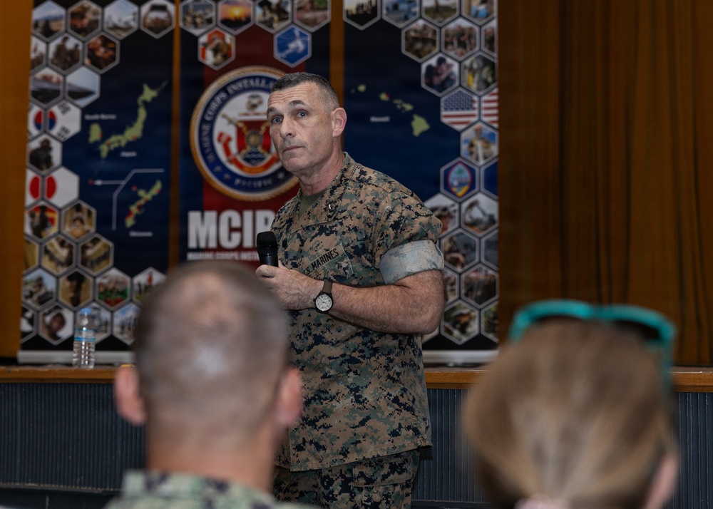 A U.S. Marine in digital camouflage (MARPAT) uniform is addressing an audience while holding a microphone. The setting appears to be an official briefing or town hall event at a Marine Corps Installations Pacific (MCIPAC) facility. An audience is visible in the foreground, seated with their backs to the camera.