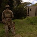 A military K-9 handler in full tactical gear stands with a military working dog, facing a deteriorating concrete structure in a vegetated outdoor environment. The handler's vest is clearly marked 'K-9 UNIT' and bears a unit patch, with a name tape reading 'STANGEL' on the hat. The scene appears to be a training exercise or patrol operation near an abandoned or mock building.