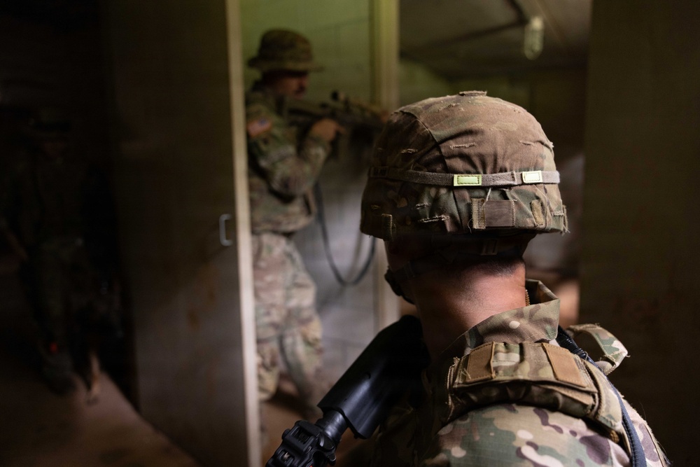 The image depicts military personnel conducting what appears to be a close-quarters battle (CQB) or urban warfare training exercise inside a building. Soldiers in MultiCam/OCP camouflage uniforms are performing a room-clearing or breach-and-entry drill through a doorway. Smoke or haze is visible in the background, consistent with training simulation conditions.
