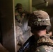 The image depicts military personnel conducting what appears to be a close-quarters battle (CQB) or urban warfare training exercise inside a building. Soldiers in MultiCam/OCP camouflage uniforms are performing a room-clearing or breach-and-entry drill through a doorway. Smoke or haze is visible in the background, consistent with training simulation conditions.