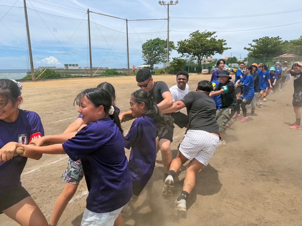 A large group of people are engaged in a tug-of-war competition on a dirt field near a coastal area. Participants wearing purple and blue shirts are pulling against each other with visible physical effort, kicking up dust from the ground. The setting appears to be an outdoor sports field, likely during a community or school event.
