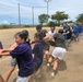A large group of people are engaged in a tug-of-war competition on a dirt field near a coastal area. Participants wearing purple and blue shirts are pulling against each other with visible physical effort, kicking up dust from the ground. The setting appears to be an outdoor sports field, likely during a community or school event.