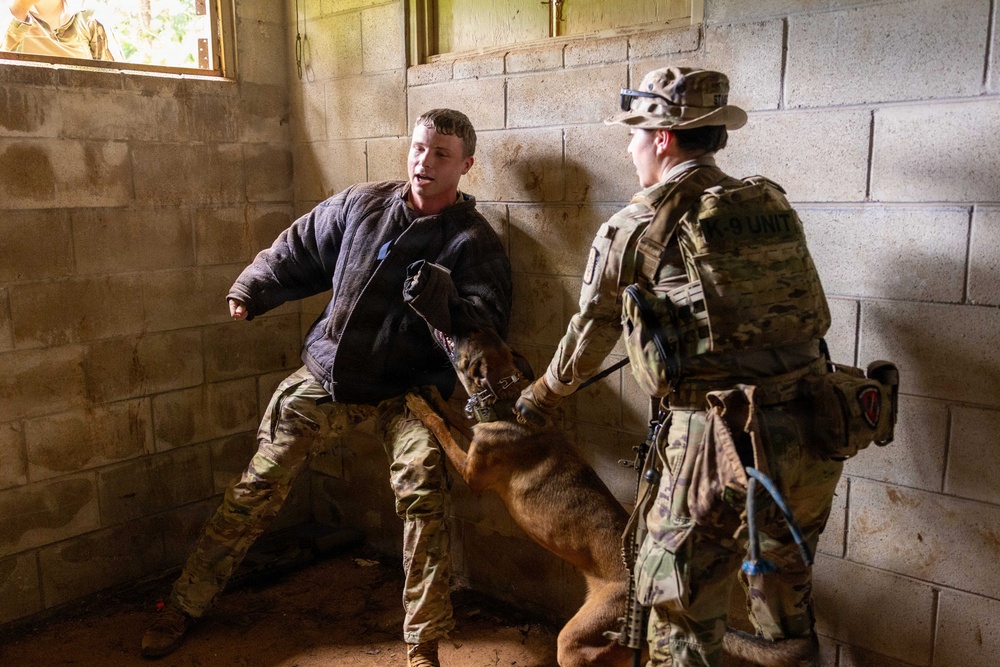 A military K-9 unit training exercise is taking place inside a concrete block structure. A soldier wearing a padded bite suit jacket is acting as a decoy/aggressor while a K-9 handler in full camouflage uniform and 'K-9 UNIT' patch controls a military working dog during an apprehension drill. The scene depicts a controlled training scenario, not an actual enforcement action.