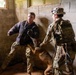 A military K-9 unit training exercise is taking place inside a concrete block structure. A soldier wearing a padded bite suit jacket is acting as a decoy/aggressor while a K-9 handler in full camouflage uniform and 'K-9 UNIT' patch controls a military working dog during an apprehension drill. The scene depicts a controlled training scenario, not an actual enforcement action.