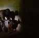 A fully equipped military or law enforcement operator in multicam/OCP camouflage uniform interacts with a military working dog (appears to be a Belgian Malinois) in a low-light, concrete block structure. The handler is leaning down toward the dog, appearing to reward or direct it, suggesting a training or operational scenario. Dramatic shadows of both the handler and dog are cast on the cinder block wall behind them.
