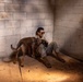 A person in military camouflage uniform sits on the floor in the corner of a concrete block structure, interacting with a medium-sized brown dog. The setting appears to be a sparse, utilitarian interior with cinder block walls and a concrete floor. Natural light enters from a window on the right side, casting dramatic shadows across the scene.