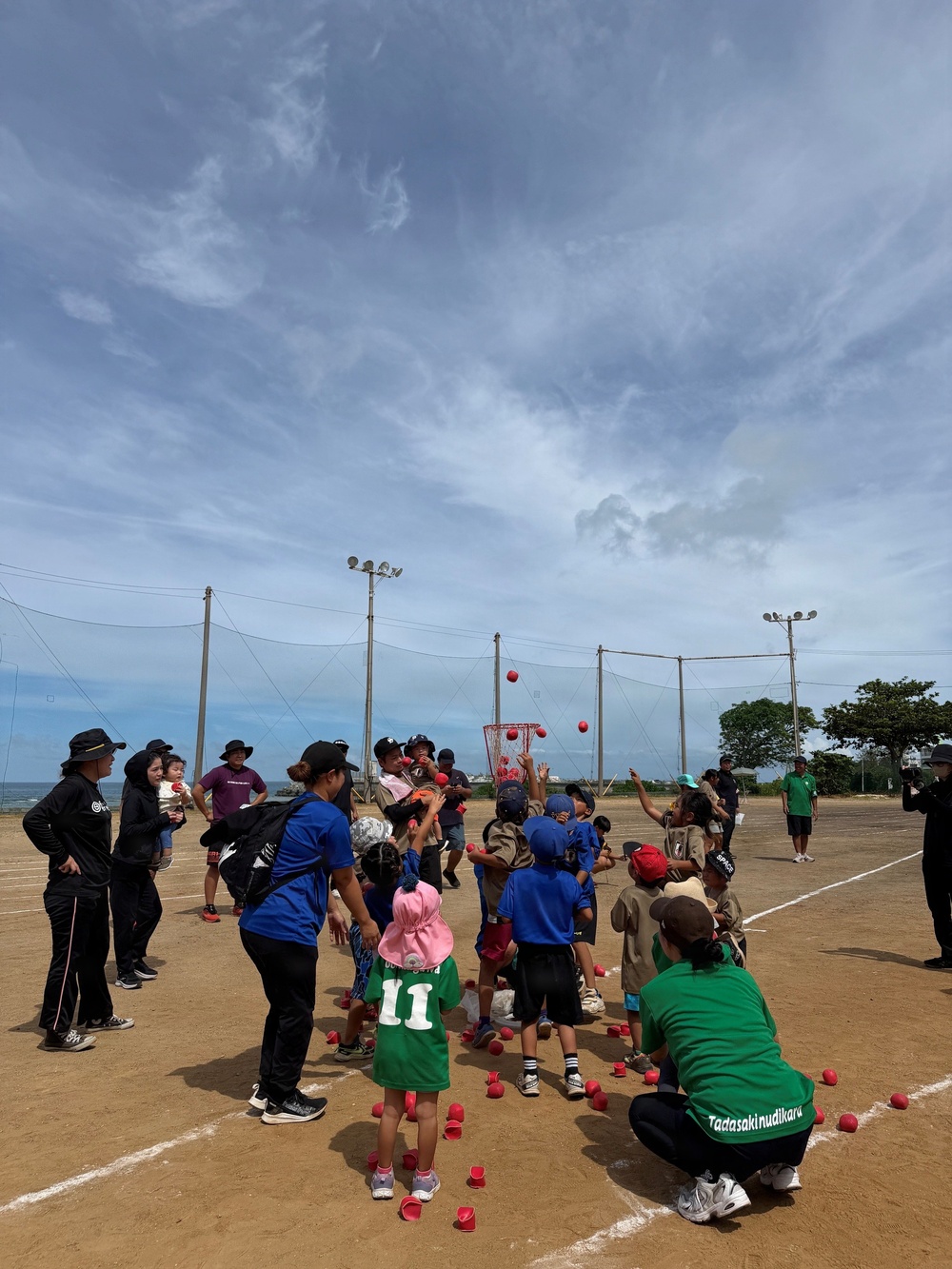 A group of children and adults are participating in an outdoor ball-tossing activity on a dirt sports field, likely a baseball or softball diamond. The children are throwing red balls toward a large basket/net target in the center, with numerous red balls scattered across the ground. The event appears to be an organized youth sports or community activity, possibly in Japan based on visible text and setting.