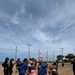 A group of children and adults are participating in an outdoor ball-tossing activity on a dirt sports field, likely a baseball or softball diamond. The children are throwing red balls toward a large basket/net target in the center, with numerous red balls scattered across the ground. The event appears to be an organized youth sports or community activity, possibly in Japan based on visible text and setting.
