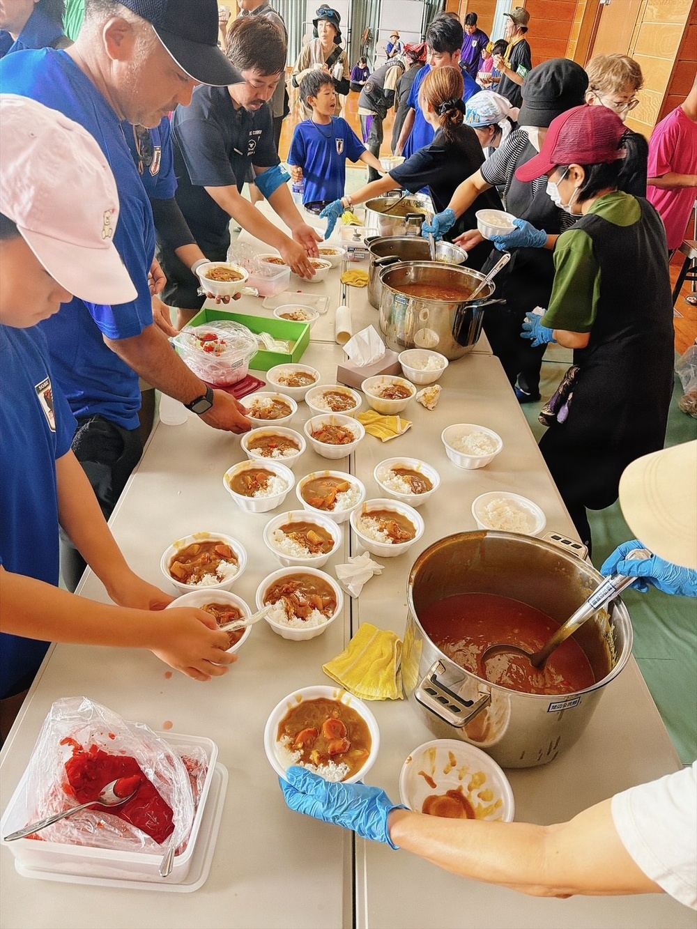 A large community food service event is taking place in what appears to be a gymnasium or community hall, where volunteers are serving Japanese curry rice (kare raisu) to a crowd of people. Multiple large stainless steel pots of curry are positioned along a long table, with numerous white disposable bowls being filled and distributed. Volunteers wearing blue gloves and various hats are actively ladling curry and rice into bowls while recipients line up to receive meals.