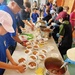 A large community food service event is taking place in what appears to be a gymnasium or community hall, where volunteers are serving Japanese curry rice (kare raisu) to a crowd of people. Multiple large stainless steel pots of curry are positioned along a long table, with numerous white disposable bowls being filled and distributed. Volunteers wearing blue gloves and various hats are actively ladling curry and rice into bowls while recipients line up to receive meals.