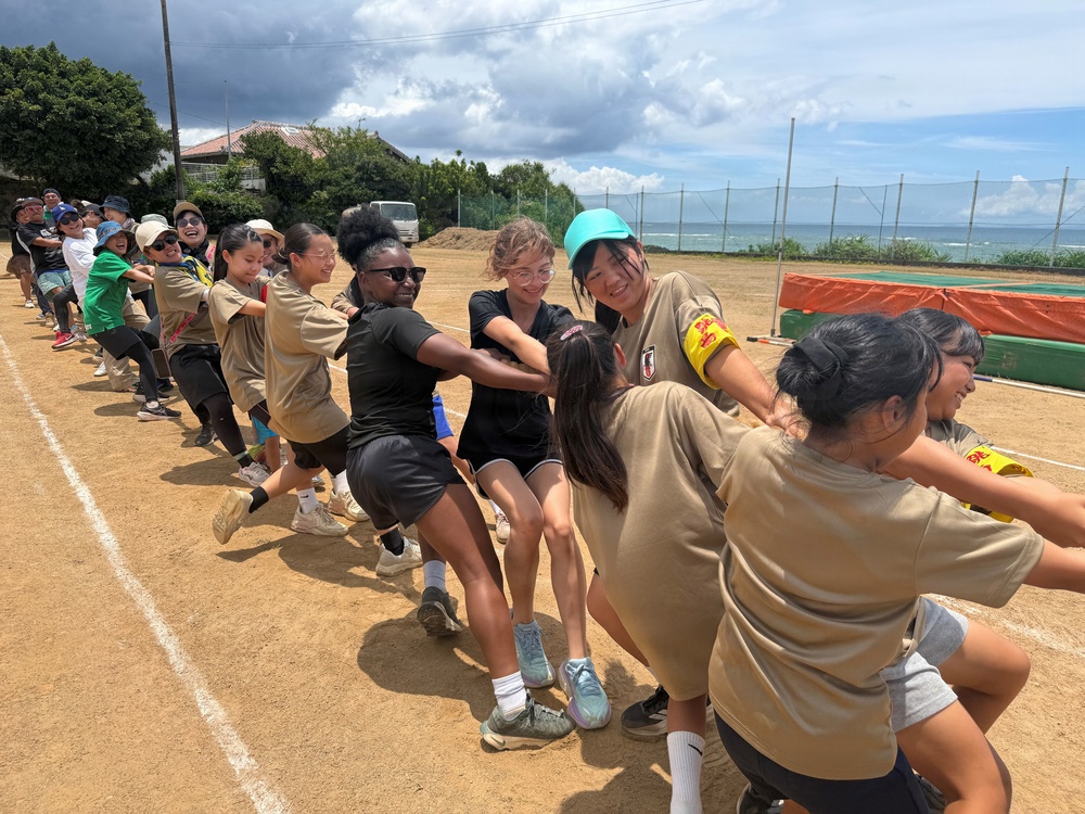 A large group of young people are participating in a tug-of-war competition on a dirt sports field near a coastal area. The participants appear to be a mixed group, some wearing tan/khaki uniforms and others in black clothing, suggesting two competing teams. The event appears to be a community or school sports day held in what looks like a coastal Japanese location.