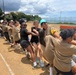 A large group of young people are participating in a tug-of-war competition on a dirt sports field near a coastal area. The participants appear to be a mixed group, some wearing tan/khaki uniforms and others in black clothing, suggesting two competing teams. The event appears to be a community or school sports day held in what looks like a coastal Japanese location.