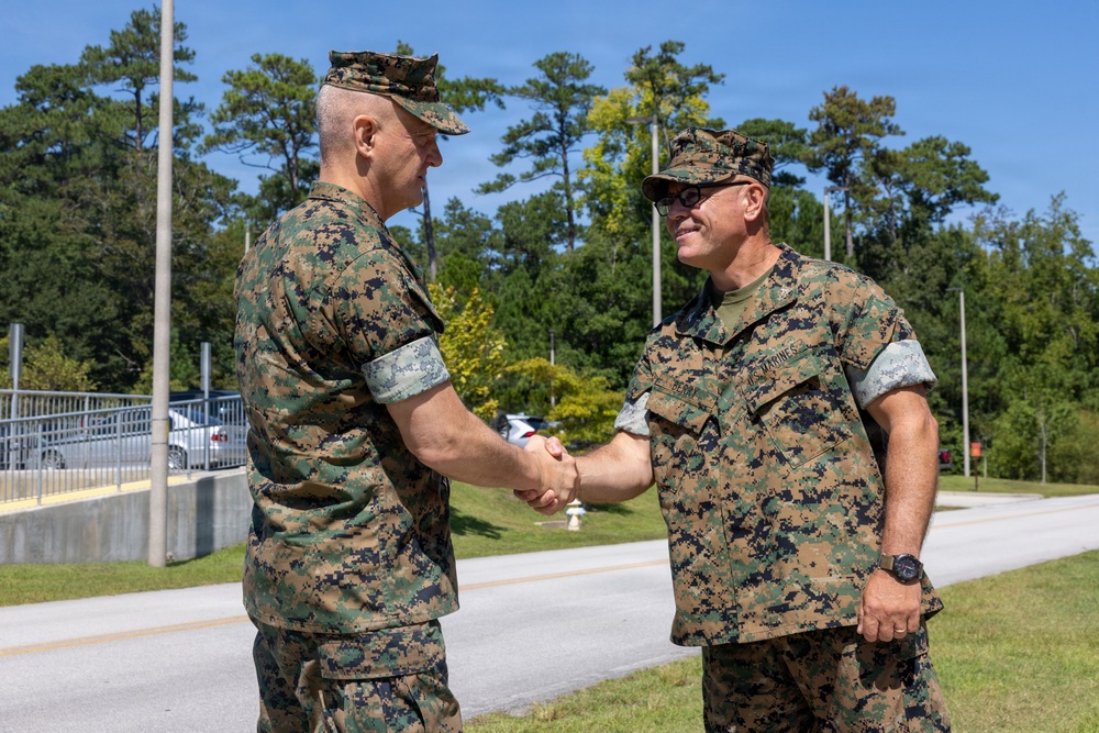 Two U.S. Marines in MARPAT woodland camouflage uniforms are shaking hands outdoors on what appears to be a military installation. Both individuals are wearing the standard Marine Corps Combat Utility Uniform (MCCUU) with covers (caps). The setting appears to be a sunny day near a road or pathway on a military base.
