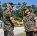 Two U.S. Marines in MARPAT woodland camouflage uniforms are shaking hands outdoors on what appears to be a military installation. Both individuals are wearing the standard Marine Corps Combat Utility Uniform (MCCUU) with covers (caps). The setting appears to be a sunny day near a road or pathway on a military base.