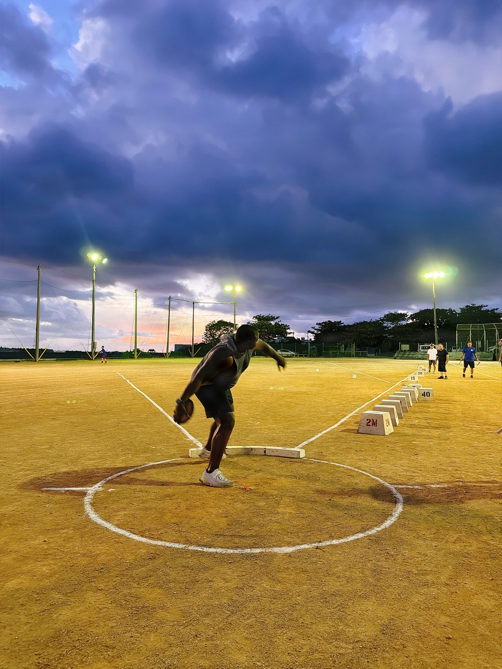 An athlete is captured mid-motion inside a throwing circle on a track and field facility, appearing to perform a discus or hammer throw during evening hours. The venue is illuminated by floodlights against a dramatic, cloud-filled twilight sky with hints of sunset color on the horizon. Several other individuals are visible in the background near distance marker blocks.