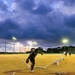 An athlete is captured mid-motion inside a throwing circle on a track and field facility, appearing to perform a discus or hammer throw during evening hours. The venue is illuminated by floodlights against a dramatic, cloud-filled twilight sky with hints of sunset color on the horizon. Several other individuals are visible in the background near distance marker blocks.