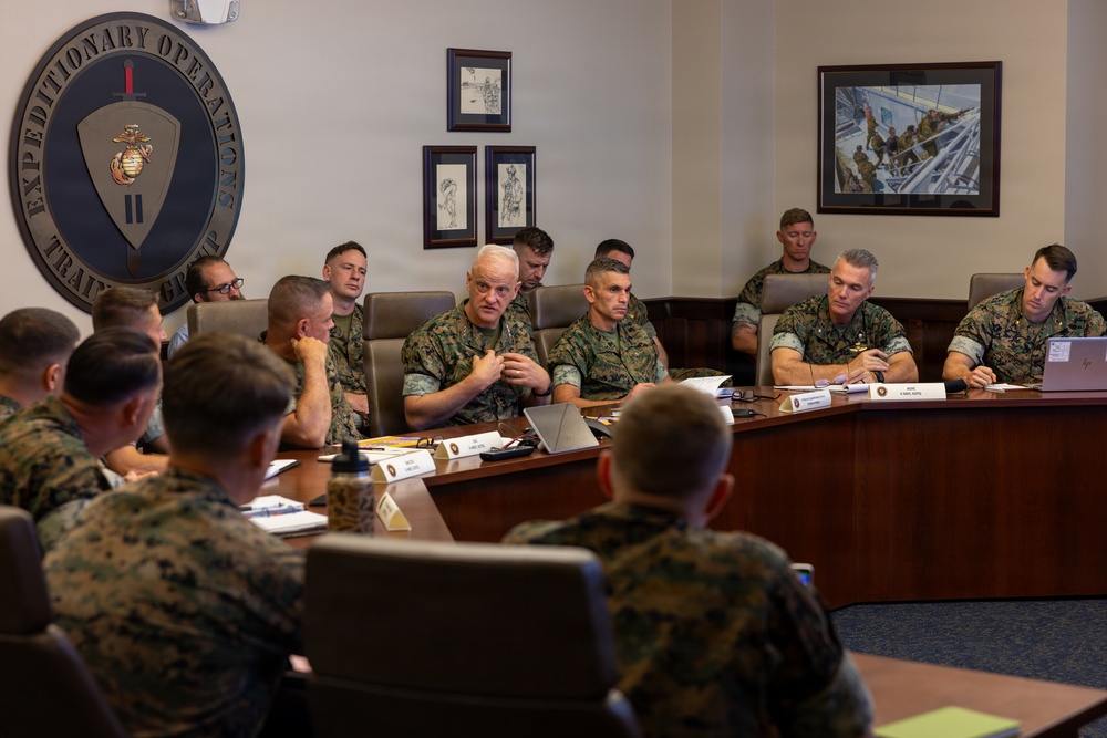 A formal military briefing or meeting is taking place in a conference room belonging to the II Marine Expeditionary Force Expeditionary Operations Training Group. Multiple Marines in woodland MARPAT camouflage uniforms are seated around a large curved conference table, with one senior officer appearing to speak or present. The room features military artwork, name placards, laptops, and the unit's official emblem on the wall.