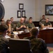 A formal military briefing or meeting is taking place in a conference room belonging to the II Marine Expeditionary Force Expeditionary Operations Training Group. Multiple Marines in woodland MARPAT camouflage uniforms are seated around a large curved conference table, with one senior officer appearing to speak or present. The room features military artwork, name placards, laptops, and the unit's official emblem on the wall.