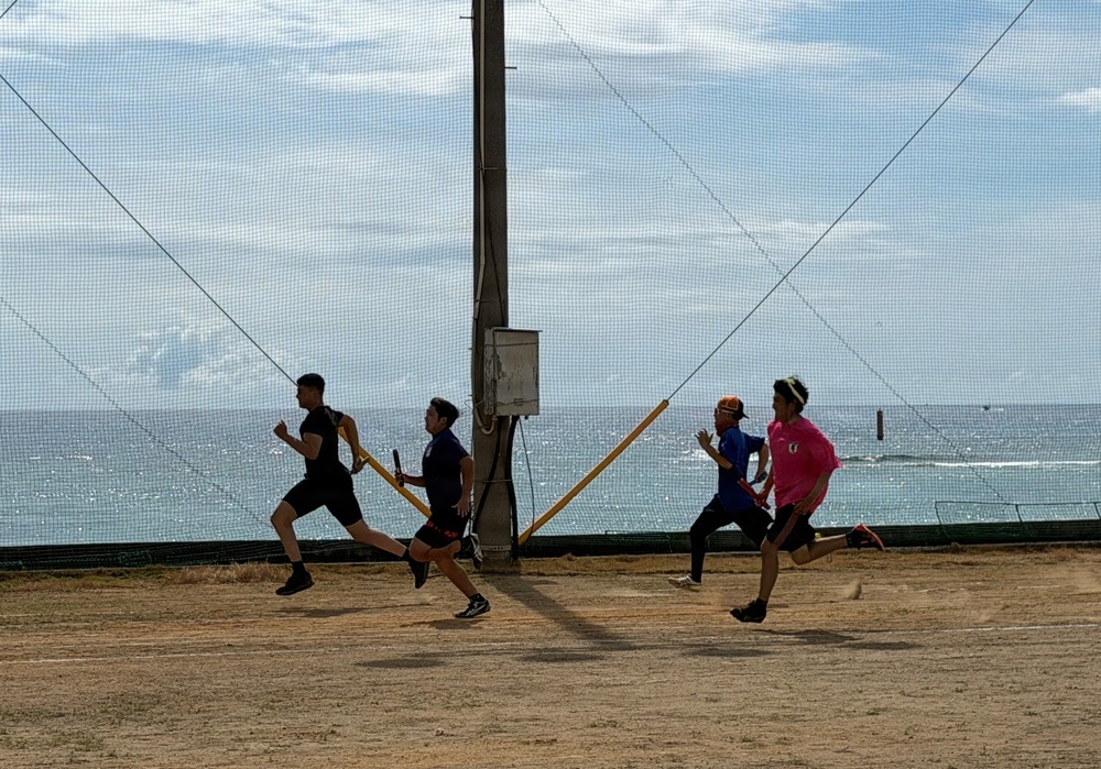 Four young males are running on a dirt/sand surface near a coastal area, appearing to participate in a relay race or sprint drill. They are running in the same direction past a central pole structure with netting, with the ocean visible in the background. The runners are wearing athletic clothing in varying colors including black, purple, blue, and pink.