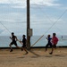 Four young males are running on a dirt/sand surface near a coastal area, appearing to participate in a relay race or sprint drill. They are running in the same direction past a central pole structure with netting, with the ocean visible in the background. The runners are wearing athletic clothing in varying colors including black, purple, blue, and pink.