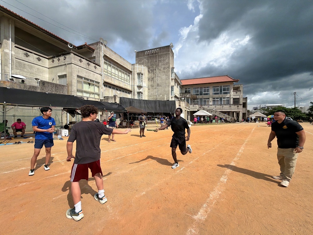 A group of people are engaged in an outdoor sports activity, likely a relay race or baton-passing drill, on a dirt track or field. The setting appears to be a schoolyard or community sports ground in an East Asian location, possibly Japan or Okinawa, based on the architectural style of the surrounding buildings. The event appears to be a community or school sports day with spectators visible in the background.