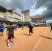A group of people are engaged in an outdoor sports activity, likely a relay race or baton-passing drill, on a dirt track or field. The setting appears to be a schoolyard or community sports ground in an East Asian location, possibly Japan or Okinawa, based on the architectural style of the surrounding buildings. The event appears to be a community or school sports day with spectators visible in the background.
