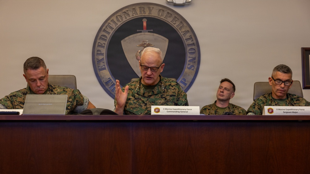 Four U.S. Marine Corps officers are seated at a conference table during an official briefing or meeting. The central figure, identified by a nameplate as the II Marine Expeditionary Force Commanding General, is speaking with a hand gesture. The setting is a formal military conference room with the II Marine Expeditionary Force Expeditionary Operations seal prominently displayed on the wall behind them.