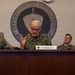 Four U.S. Marine Corps officers are seated at a conference table during an official briefing or meeting. The central figure, identified by a nameplate as the II Marine Expeditionary Force Commanding General, is speaking with a hand gesture. The setting is a formal military conference room with the II Marine Expeditionary Force Expeditionary Operations seal prominently displayed on the wall behind them.
