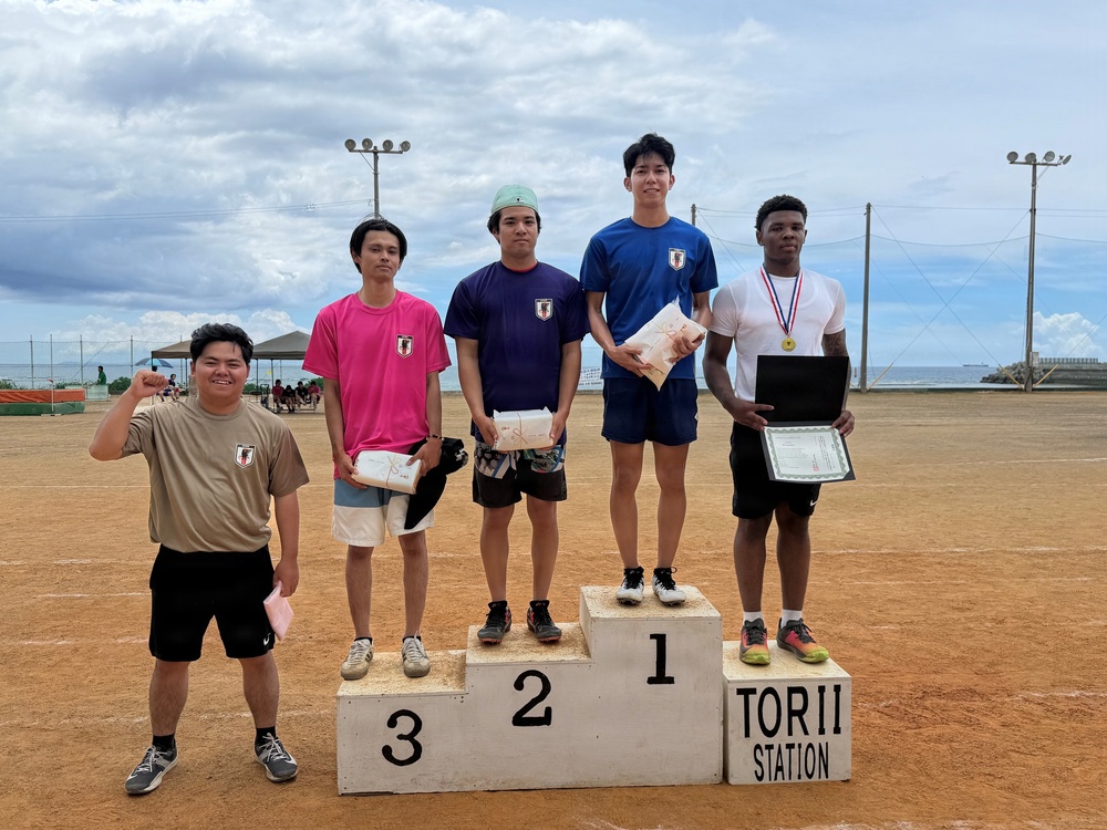 A podium ceremony at Torii Station featuring five young male athletes standing on or beside a three-tiered awards podium. The first-place finisher on the right wears a medal and holds a certificate, while second and third place holders carry wrapped packages. A fifth individual stands off the podium to the left, raising a fist in celebration.