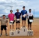A podium ceremony at Torii Station featuring five young male athletes standing on or beside a three-tiered awards podium. The first-place finisher on the right wears a medal and holds a certificate, while second and third place holders carry wrapped packages. A fifth individual stands off the podium to the left, raising a fist in celebration.