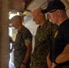 Four individuals are standing in what appears to be a military field environment, possibly inside a tent or temporary structure. Two individuals are wearing U.S. Marines MARPAT camouflage uniforms, while two others are wearing black tactical shirts with a yellow emblem. All four appear to have their heads bowed, suggesting a moment of prayer, reflection, or a briefing.