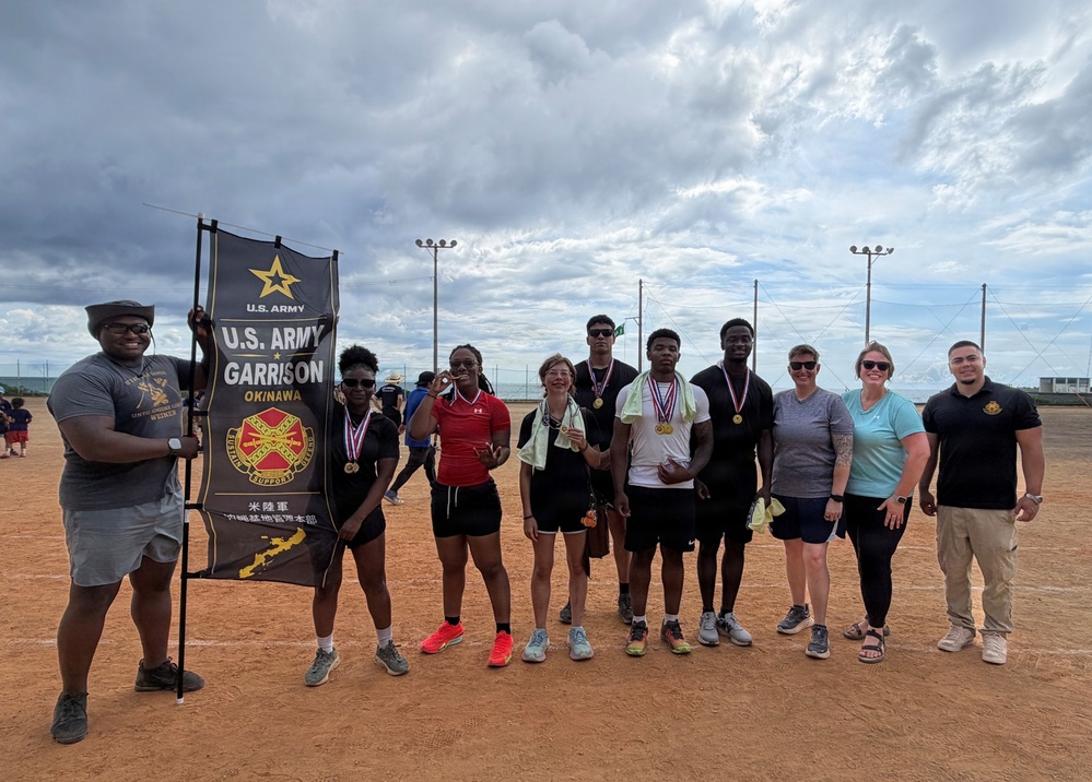 A group of approximately ten people pose together on a dirt sports field, several wearing medals around their necks. They are gathered under a U.S. Army Garrison Okinawa banner, suggesting a military athletic competition or sports event. The setting appears to be an outdoor softball or baseball field with light poles visible in the background.
