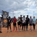 A group of approximately ten people pose together on a dirt sports field, several wearing medals around their necks. They are gathered under a U.S. Army Garrison Okinawa banner, suggesting a military athletic competition or sports event. The setting appears to be an outdoor softball or baseball field with light poles visible in the background.