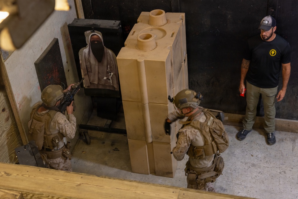 Two military personnel in desert camouflage uniforms and tactical gear are conducting a close-quarters battle (CQB) training exercise at an indoor shooting range or training facility. They are engaging a paper/cardboard target depicting a hooded figure, with large modular foam/rubber training barriers used as cover. A civilian instructor in a black t-shirt and cap observes the drill from the side.