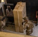 Two military personnel in desert camouflage uniforms and tactical gear are conducting a close-quarters battle (CQB) training exercise at an indoor shooting range or training facility. They are engaging a paper/cardboard target depicting a hooded figure, with large modular foam/rubber training barriers used as cover. A civilian instructor in a black t-shirt and cap observes the drill from the side.