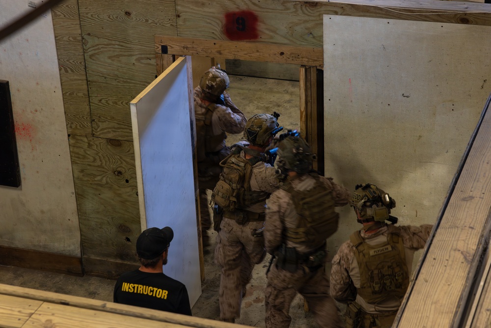 Military or law enforcement personnel in full tactical gear are conducting a room-clearing or close-quarters battle (CQB) training exercise inside a mock building structure. A civilian instructor wearing a black shirt labeled 'INSTRUCTOR' observes the drill from a lower vantage point. The facility appears to be an indoor shoot house or training compound with numbered rooms and plywood construction.