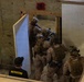 Military or law enforcement personnel in full tactical gear are conducting a room-clearing or close-quarters battle (CQB) training exercise inside a mock building structure. A civilian instructor wearing a black shirt labeled 'INSTRUCTOR' observes the drill from a lower vantage point. The facility appears to be an indoor shoot house or training compound with numbered rooms and plywood construction.