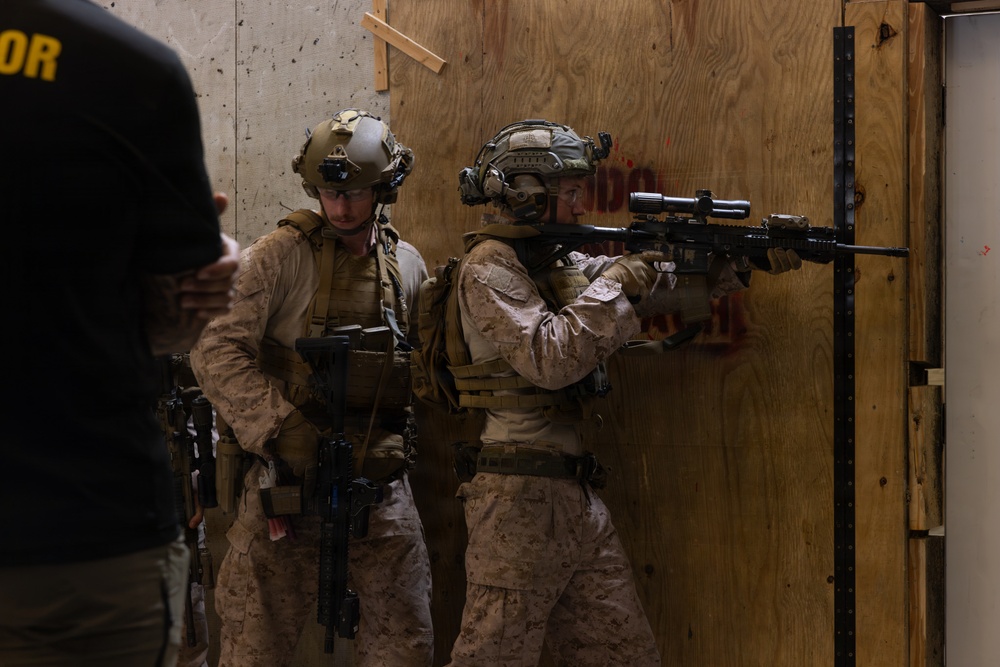 Two fully equipped military personnel in desert MARPAT camouflage uniforms are conducting what appears to be a close-quarters battle (CQB) training exercise inside a simulated building structure with plywood walls. One operator is aiming a rifle with a scope toward a doorway while the other stands in a support position. A third individual in civilian/instructor attire is partially visible on the left, suggesting an observed training scenario.