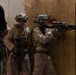 Two fully equipped military personnel in desert MARPAT camouflage uniforms are conducting what appears to be a close-quarters battle (CQB) training exercise inside a simulated building structure with plywood walls. One operator is aiming a rifle with a scope toward a doorway while the other stands in a support position. A third individual in civilian/instructor attire is partially visible on the left, suggesting an observed training scenario.