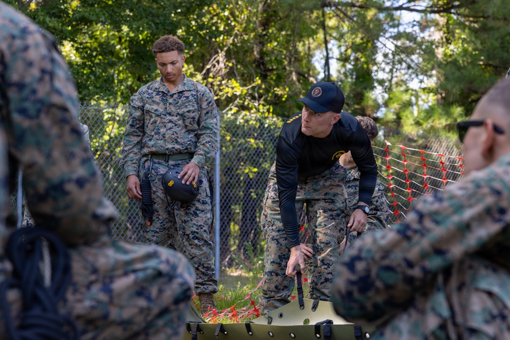 A group of military personnel in MARPAT camouflage uniforms are engaged in a training exercise outdoors. A central figure wearing a black long-sleeve shirt and a dark baseball cap appears to be an instructor demonstrating a technique over a green military litter/stretcher. The setting is a wooded area with a chain-link fence and orange safety netting visible in the background.
