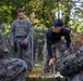 A group of military personnel in MARPAT camouflage uniforms are engaged in a training exercise outdoors. A central figure wearing a black long-sleeve shirt and a dark baseball cap appears to be an instructor demonstrating a technique over a green military litter/stretcher. The setting is a wooded area with a chain-link fence and orange safety netting visible in the background.