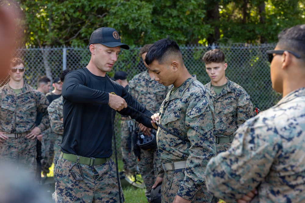 A military instructor in black civilian-style clothing and a black cap is demonstrating or inspecting equipment on a U.S. Marine in digital camouflage (MARPAT) uniform. The setting appears to be an outdoor training area with a chain-link fence and trees in the background. A group of additional Marines in uniform observe the instruction.