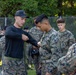 A military instructor in black civilian-style clothing and a black cap is demonstrating or inspecting equipment on a U.S. Marine in digital camouflage (MARPAT) uniform. The setting appears to be an outdoor training area with a chain-link fence and trees in the background. A group of additional Marines in uniform observe the instruction.